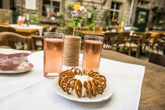 Zwei Gläser mit Weinschorle und ein Teller mit Spundekäs und Salzbrezeln stehen auf einem Tisch in einem gemütlichen Innenhof einer Straußwirtschaft., © Achim Meurer Zwei Gläser und ein Teller mit Dip und Brezeln stehen auf einem Tisch