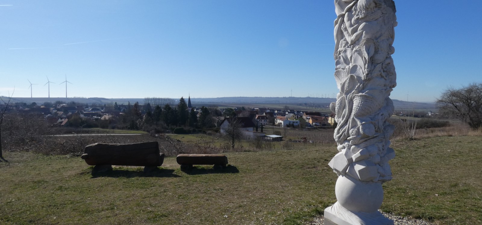 Dorn-Dürkheim, Evolutionssäule, © TSC Rhein-Selz Weiße, abstrakte Steinskulptur auf einer Wiese mit Blick auf ein Dorf und Windräder am Horizont unter klarem Himmel