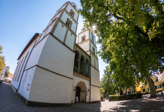 Heidenturmkirche in Guntersblum, © inMEDIA Weißes historisches Kirchengebäude mit zwei Türmen, davor ein Platz und große Bäume