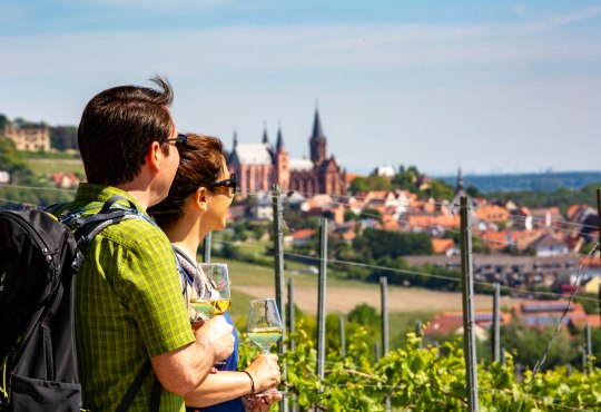 Ein Paar macht eine Pause bei einer Wanderung und blickt auf Oppenheim mit der Katharinenkirche im Hintergrund., © inMEDIA Paar mit Weingläsern blickt auf eine Stadt mit einer großen Kirche und Weinbergen im Vordergrund
