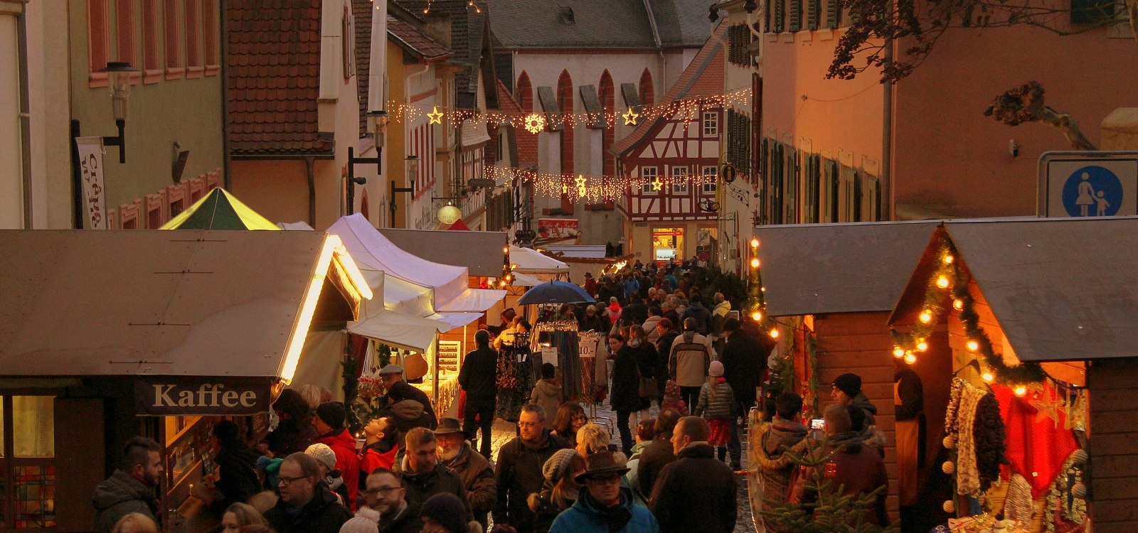 Der Oppenheimer Weihnachtsmarkt lädt mit zahlreichen Ständen die Besucher zu einem Besuch ein., © Stadt Oppenheim Belebter Weihnachtsmarkt in einer Altstadtstraße mit beleuchteten Ständen, Menschen und festlicher Dekoration aus Lichterketten und Sternen.