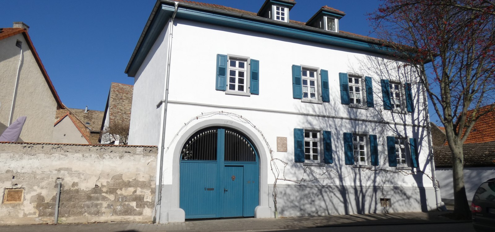 Selzen, © TSC Rhein-Selz Weißes zweistöckiges Haus mit blauen Fensterläden und großem blauem Tor unter blauem Himmel