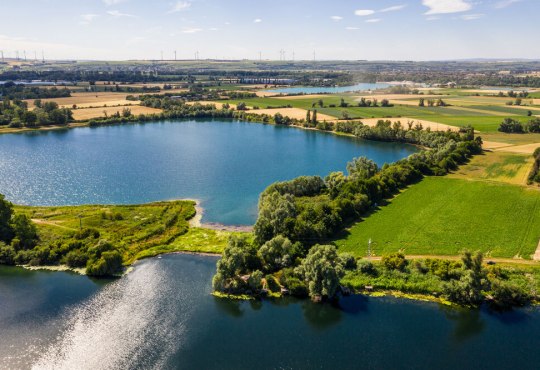 Landschaft mit zwei verbundenen Seen, umgeben von grünen Feldern, Bäumen und Windrädern am Horizont unter blauem Himmel