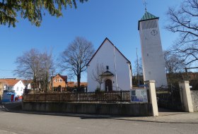 Weiße Kirche mit spitzem Dach und angrenzendem Turm mit Uhr in Ortsmitte