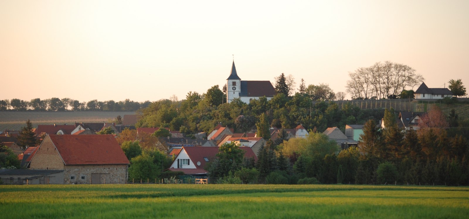 Blick auf Dorf mit einer Kirche, im Vordergrund gr&uuml;ne Felder bei Sonnenuntergang