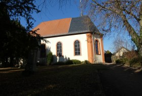 Kleine Kirche mit wei&szlig;em Putz und orangefarbenen Fensterrahmen, umgeben von B&auml;umen und Schatten am sonnigen Tag
