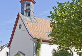 Wei&szlig;e Kirche mit rotem Dach und kleinem Turm, umgeben von B&auml;umen und Menschen vor dem Eingang