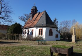 Kleine Kirche umgeben von B&auml;umen und einer Bank im Vordergrund