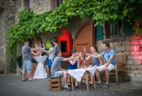 Menschen sitzen drau&szlig;en vor einem Kelterhaus und sto&szlig;en mit Gl&auml;sern an