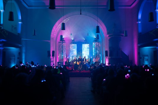 Musiker spielen auf einer Bühne in einer Kirche mit bunten Lichteffekten, Publikum trägt leuchtende Kopfhörer