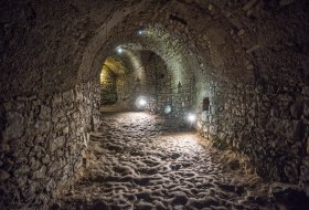 Beleuchteter unterirdischer Gang aus grobem Stein mit unebenem Boden 