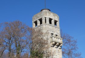 Steinerner Turm, umgeben von B&auml;umen und Hecken unter klarem blauem Himmel