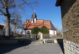 Kleine Kirche mit rotem Dach und kleinem Turm, umgeben von B&auml;umen und einer Mauer mit Tor