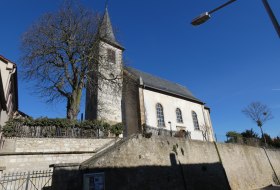 Wei&szlig;e Kirche mit Turm und spitzem Dach vor klarem blauem Himmel, im Vordergrund eine Steinmauer und ein kahler Baum