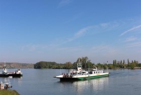 F&auml;hre auf einem Fluss bei klarem Himmel, Ufer mit Gras und B&auml;umen im Hintergrund