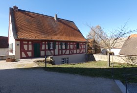 Ein Fachwerkhaus mit rotem Dach und gr&uuml;ner T&uuml;r, umgeben von Wiese und B&auml;umen.