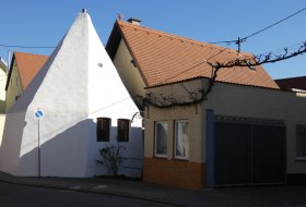 Wei&szlig;es, dreieckiges Geb&auml;ude neben einem Haus mit rotem Ziegeldach an einer Stra&szlig;enecke.