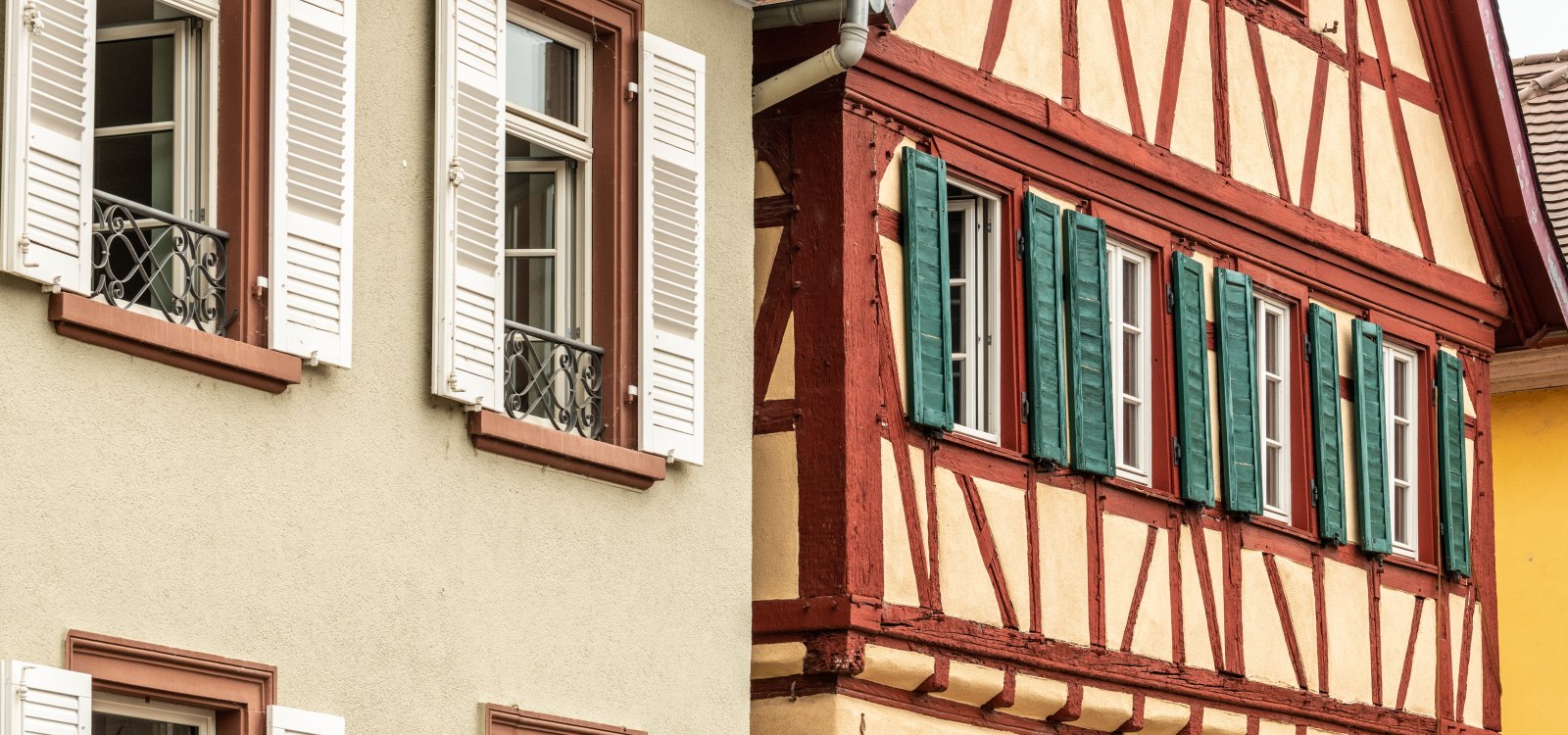 Fachwerkhaus mit gr&uuml;nen Fensterl&auml;den neben einem hellen Geb&auml;ude mit wei&szlig;en Fensterl&auml;den