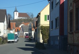 Stra&szlig;e in einem Dorf mit H&auml;usern, im Hintergrund eine Kirche