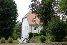Wei&szlig;es zweist&ouml;ckiges Haus mit rotem Satteldach, umgeben von gr&uuml;nen B&auml;umen und Str&auml;uchern