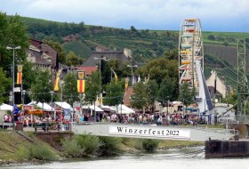 Weinfest am Flussufer mit Menschen, St&auml;nden und Riesenrad. Im Hintergrund sind Weinberge. 