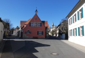 Stra&szlig;e in einem Dorf mit einem roten Fachwerkhaus in der Mitte und weiteren H&auml;usern mit wei&szlig;en Fassaden und gr&uuml;nen Fensterl&auml;den bei klarem Himmel