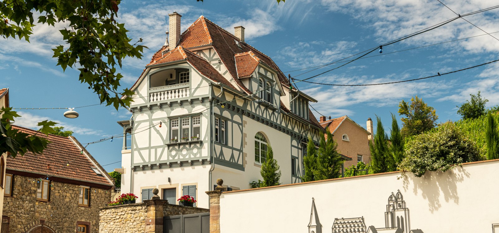 Fachwerkhaus mit roten Ziegeldach und Blumenkästen, umgeben von Bäumen und einer weißen Mauer mit Zeichnung von Gebäuden