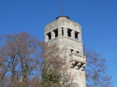 Steinerner Turm, umgeben von Bäumen und Hecken unter klarem blauem Himmel