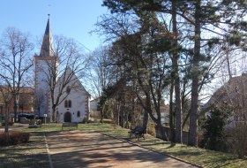 Kirche mit spitzem Turm neben einem von B&auml;umen ges&auml;umten Weg mit Parkb&auml;nken