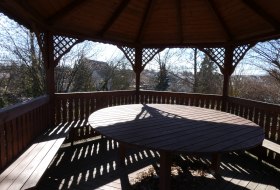 Holzpavillon mit Tisch und Sitzb&auml;nken, umgeben von B&auml;umen und H&auml;usern im Hintergrund