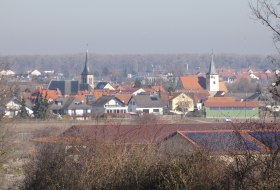 Blick auf ein Dorf mit zwei Kircht&uuml;rmen, im Vordergrund Felder und B&auml;ume