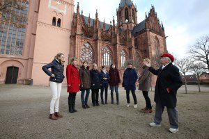 Eine Gruppe steht vor einer gotischen Kirche und hört einer Gästeführerin aufmerksam zu