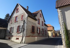 Steinernes Wohnhaus mit roten Fensterrahmen an einer ruhigen Stra&szlig;e unter blauem Himmel