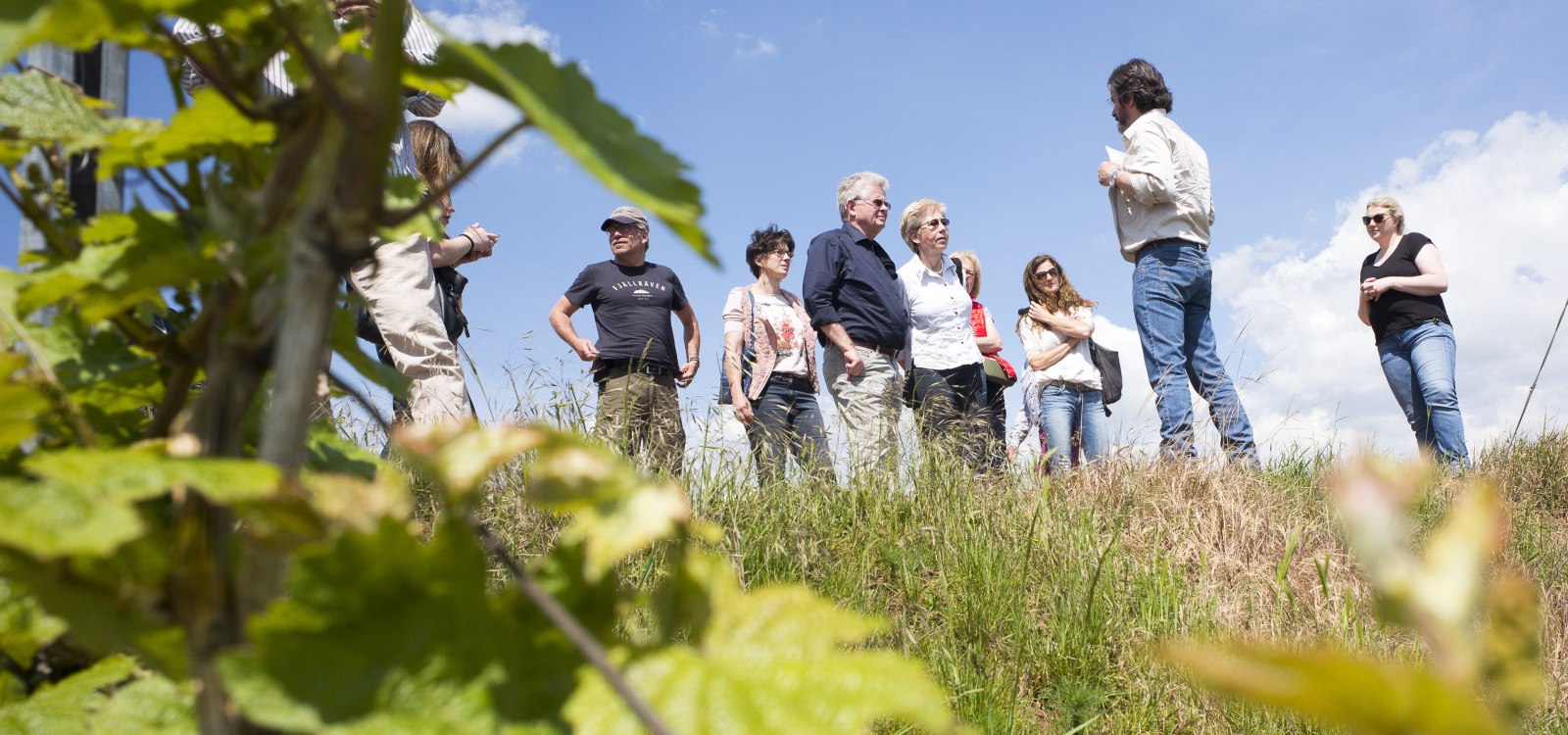 Gruppe von acht Menschen steht auf einem grasbewachsenen H&uuml;gel unter blauem Himmel, eine Person spricht zur Gruppe