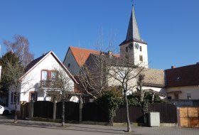 Kirchturm mit Uhr hinter Wohnh&auml;usern und kahlen B&auml;umen bei klarem Himmel