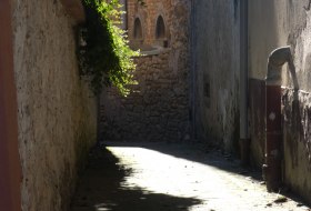 Sonnendurchflutete enge Gasse mit alten Steinmauern und Fenstern, teilweise von Pflanzen beschattet