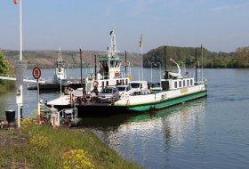F&auml;hre mit mehreren Fahrzeugen auf einem Fluss, umgeben von gr&uuml;ner Landschaft und H&uuml;geln im Hintergrund