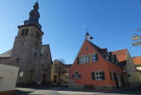 Historische Kirche mit Turm neben einem roten Fachwerkhaus mit blauen Fensterl&auml;den unter klarem blauem Himmel