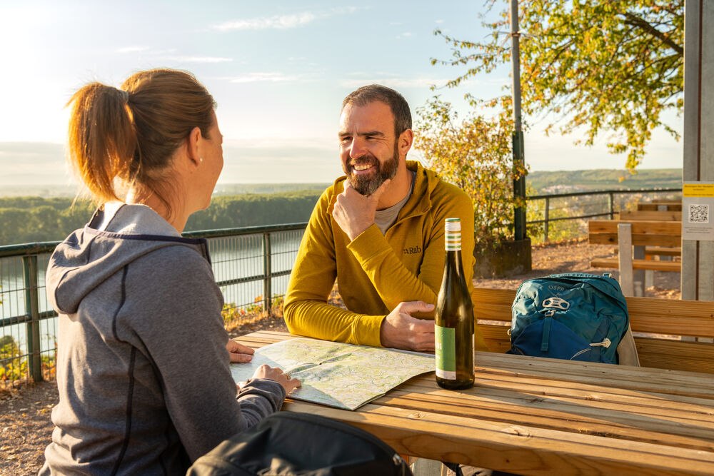 Ein Paar sitzt an einem Tisch im Freien und betrachtet eine Wanderkarte. Im Hintergrund sind Weinberge und ein Fluss zu sehen.