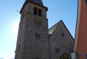Kirchturm aus Stein mit Uhr und spitzem Dach vor blauem Himmel, daneben ein Geb&auml;ude mit orangefarbener Fassade