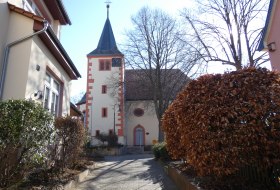 Kirche mit Kirchturm, umgeben von H&auml;usern, B&auml;umen und Str&auml;uchern an einem sonnigen Tag
