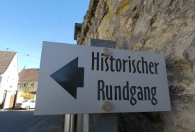 Schild mit schwarzem Pfeil nach links und der Aufschrift 'Historischer Rundgang' an einer Steinmauer