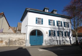 Wei&szlig;es zweist&ouml;ckiges Haus mit blauen Fensterl&auml;den und gro&szlig;em blauem Tor unter blauem Himmel