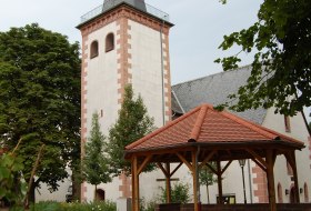Kirchturm mit spitzem Dach und wei&szlig;er Fassade mit roten Ziersteinen, davor ein h&ouml;lzerner Pavillon