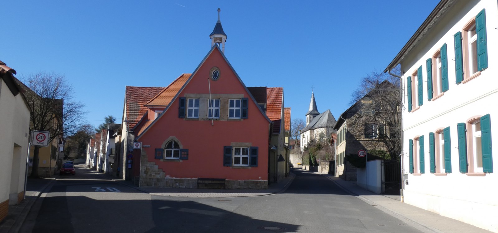 Stra&szlig;e in einem Dorf mit einem roten Fachwerkhaus in der Mitte und weiteren H&auml;usern mit wei&szlig;en Fassaden und gr&uuml;nen Fensterl&auml;den bei klarem Himmel
