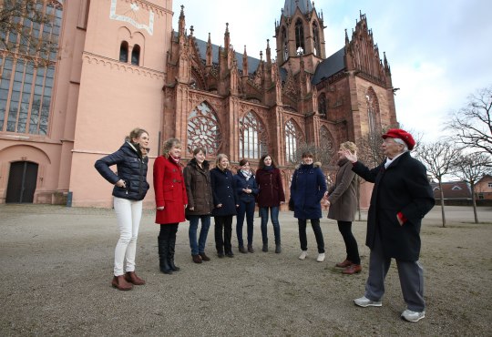 Eine Gruppe steht vor einer gotischen Kirche und hört einer Gästeführerin aufmerksam zu