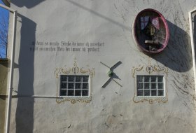 Wei&szlig;e Hauswand mit zwei kleinen Fenstern, einer Sonnenuhr und einem runden, roten Fenster mit Pflanzen. Text in deutscher Sprache an der Wand.