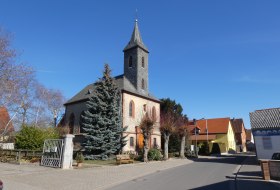 Kirche mit spitzem Turm und roten Fensterb&ouml;gen in einem ruhigen Dorf bei klarem Himmel