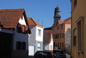 Blick auf eine ruhige Stra&szlig;e mit H&auml;usern in verschiedenen Farben und einem Kirchturm im Hintergrund.